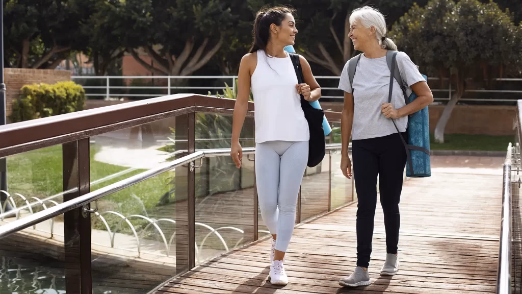 Duas mulheres caminhando e conversando ao ar livre, com tapetes de yoga a tiracolo